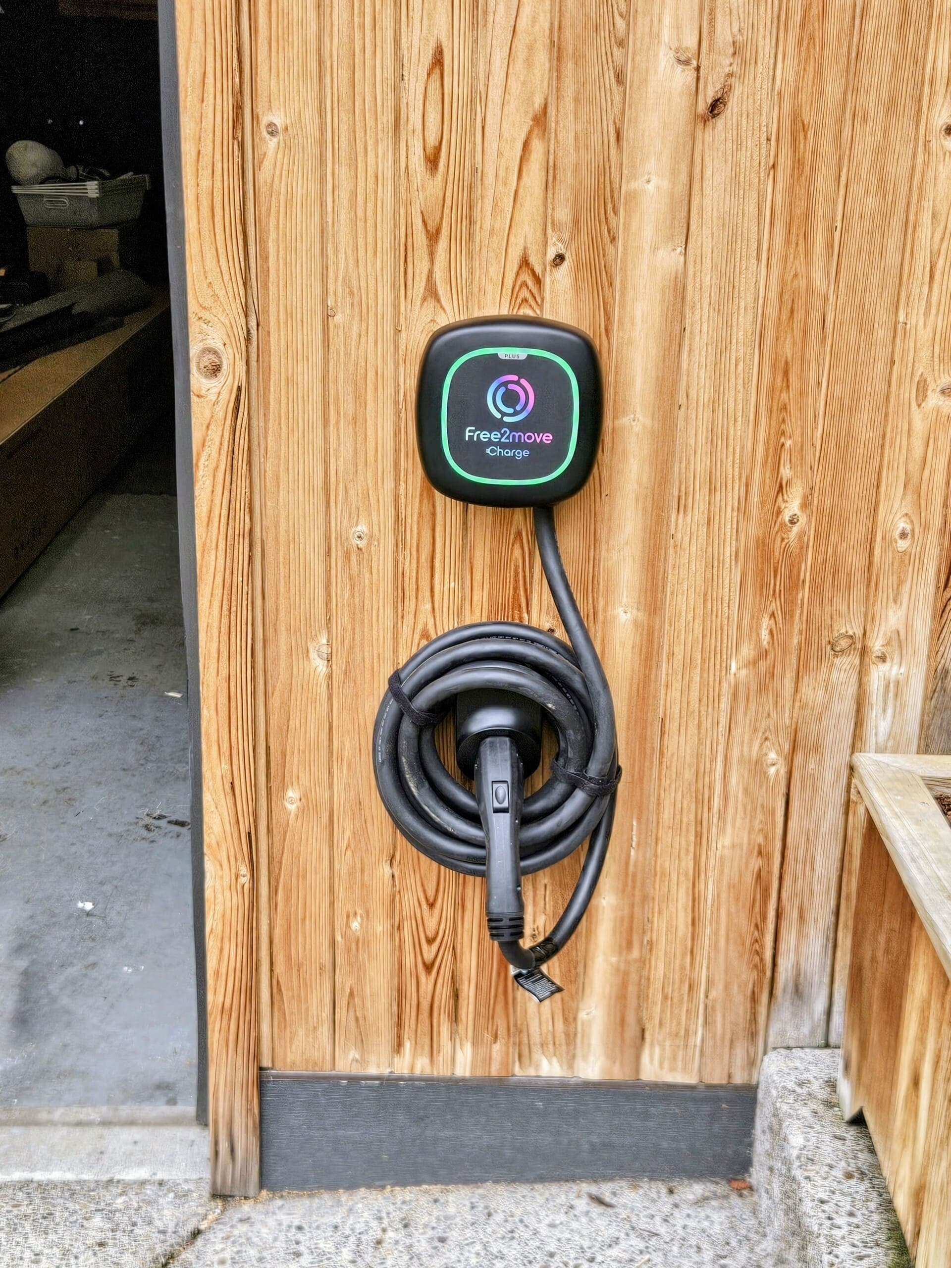 A wall-mounted EV charger installed on cedar siding beside a garage door, its cable coiled and hung
