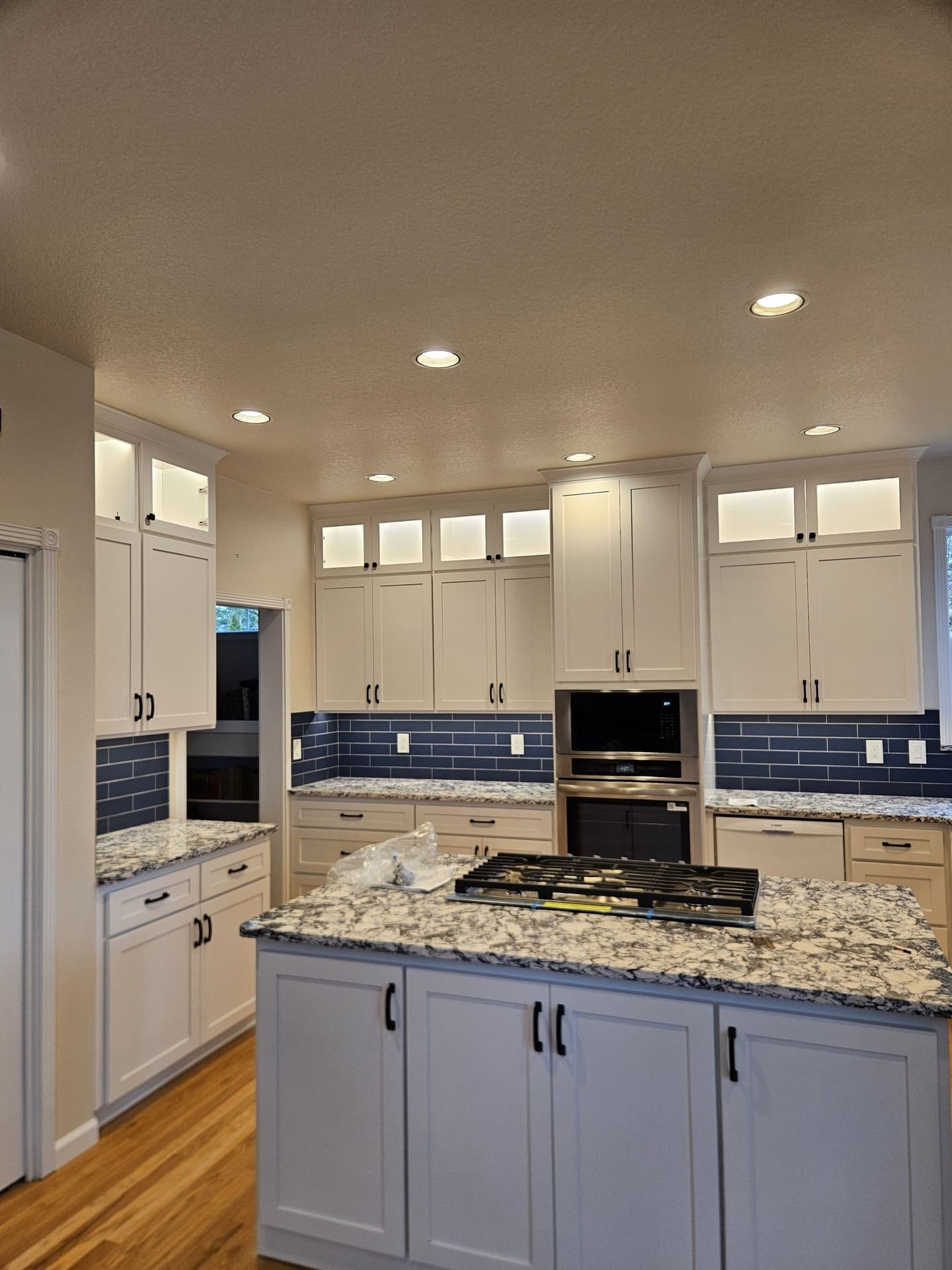 Recessed ceiling lights and lit glass front upper cabinets in a white kitchen with a blue tile backsplash