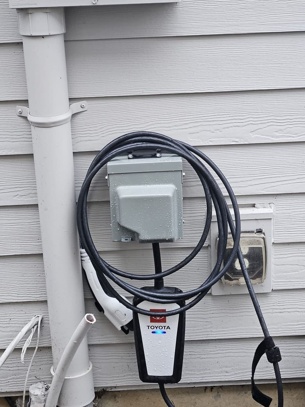 An EV charger and its weatherproof disconnect mounted on lap siding beside a downspout